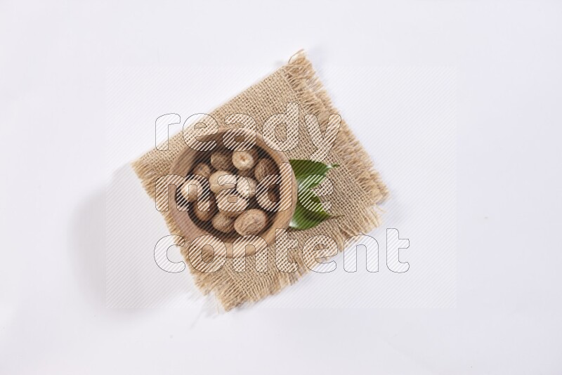 A wooden bowl full of whole nutmeg seeds on burlap fabric on a white flooring
