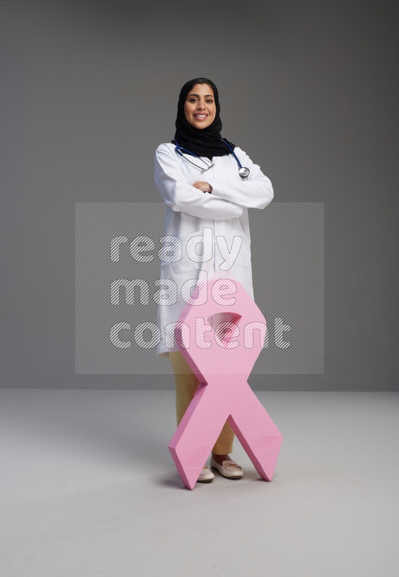 Saudi woman wearing lab coat with stethoscope standing holding awareness ribbon symbol standing on Gray background