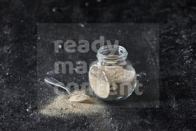 Herbal glass jar and metal spoon full of white pepper powder on textured black flooring