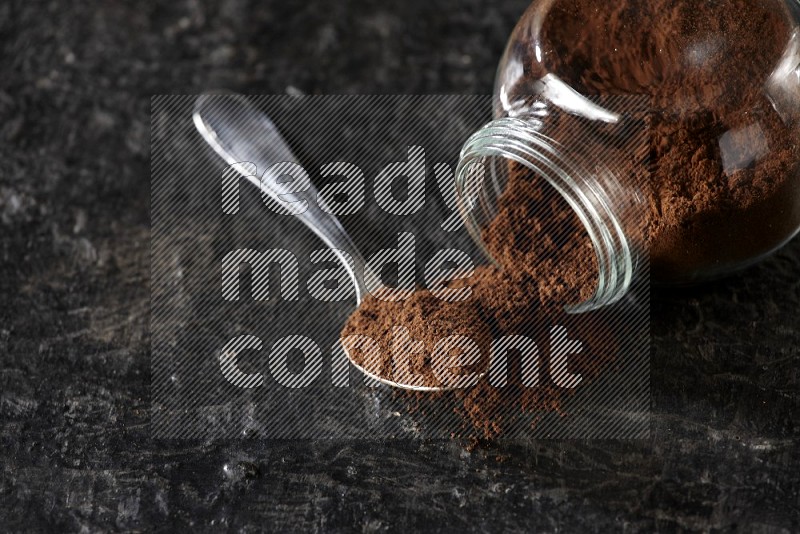 A flipped glass spice jar and a metal spoon full of cloves powder on textured black flooring