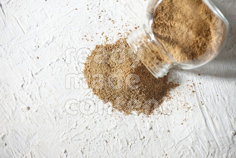 A flipped glass spice jar full of cumin powder and powder spilled out on textured white flooring