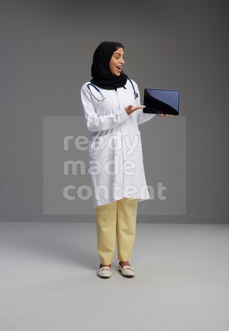 Saudi woman wearing lab coat with stethoscope standing showing tablet to camera with sign in the back on Gray background