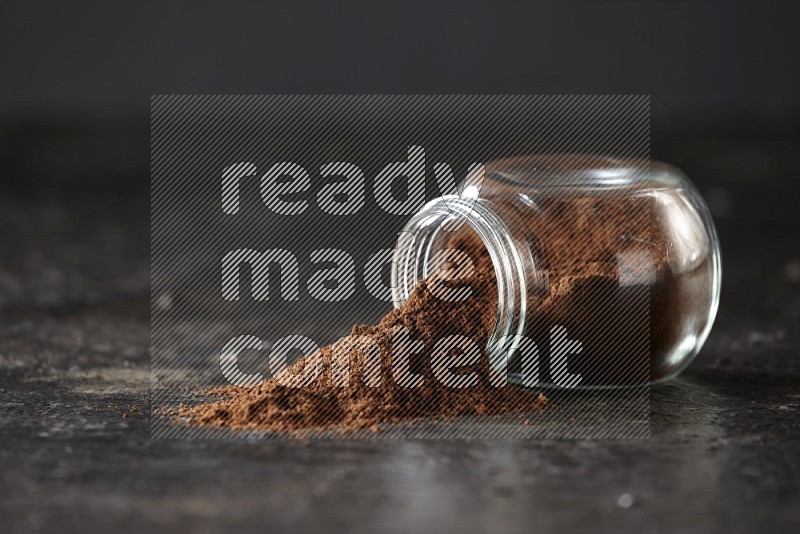 A flipped glass spice jar full of cloves powder and powder came out of it on textured black flooring