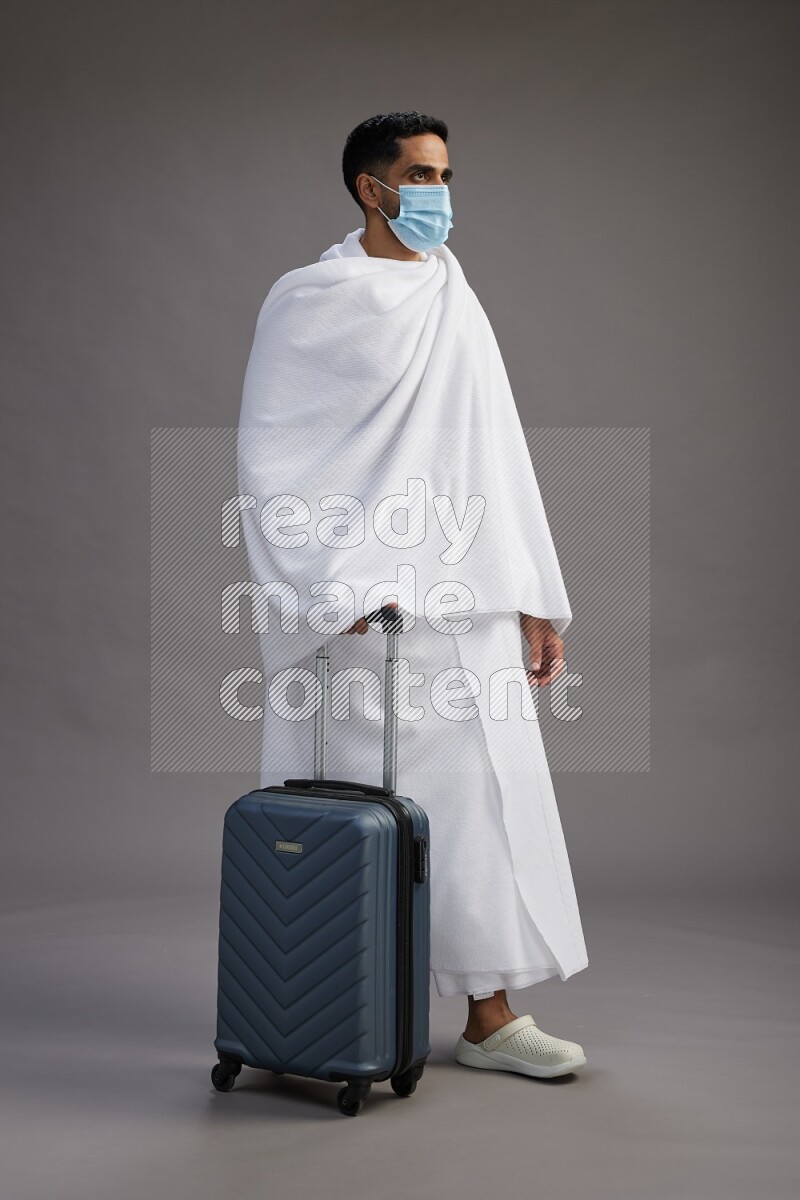 A man wearing Ehram with face mask Standing holding traveling bag on gray background