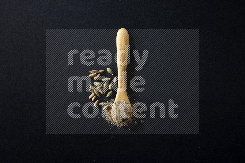 A wooden spoon full of cardamom powder and cardamom seeds beside it on black flooring