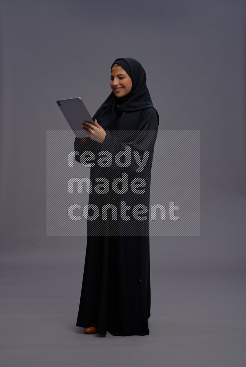 Saudi woman wearing Abaya standing working on tablet on gray background