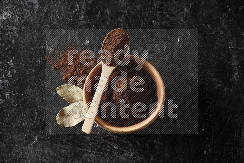 A wooden bowl and a wooden spoon full of cloves powder with laurel leaves on a textured black flooring