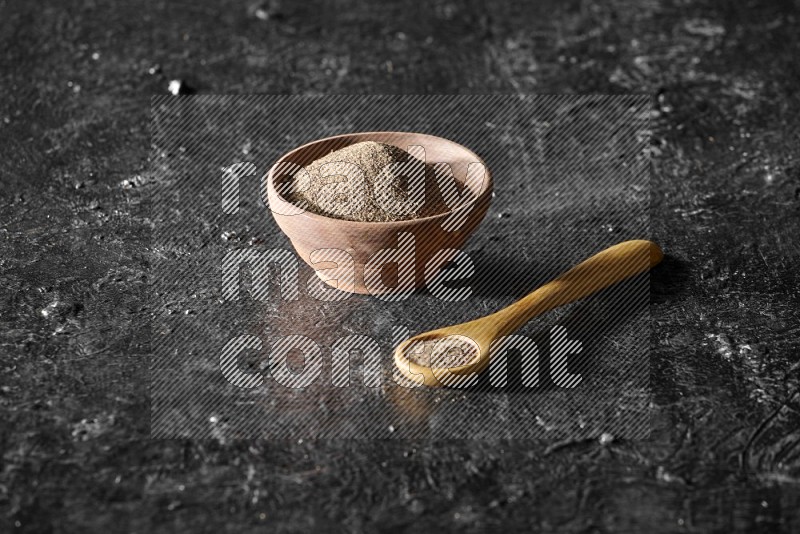 A wooden bowl and wooden spoon full of black pepper powder on a textured black flooring