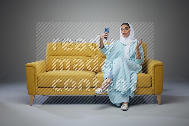 A Saudi woman wearing a light blue Abaya and white head scarf sitting on a yellow sofa and taking selfies with her phone eye level on a grey background