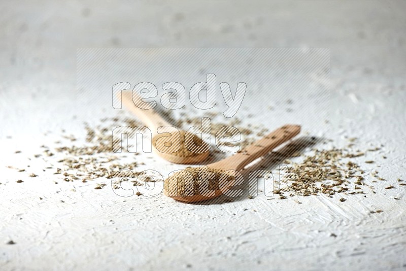 2 Wooden spoons full of cumin powder and cumin seeds on textured white flooring