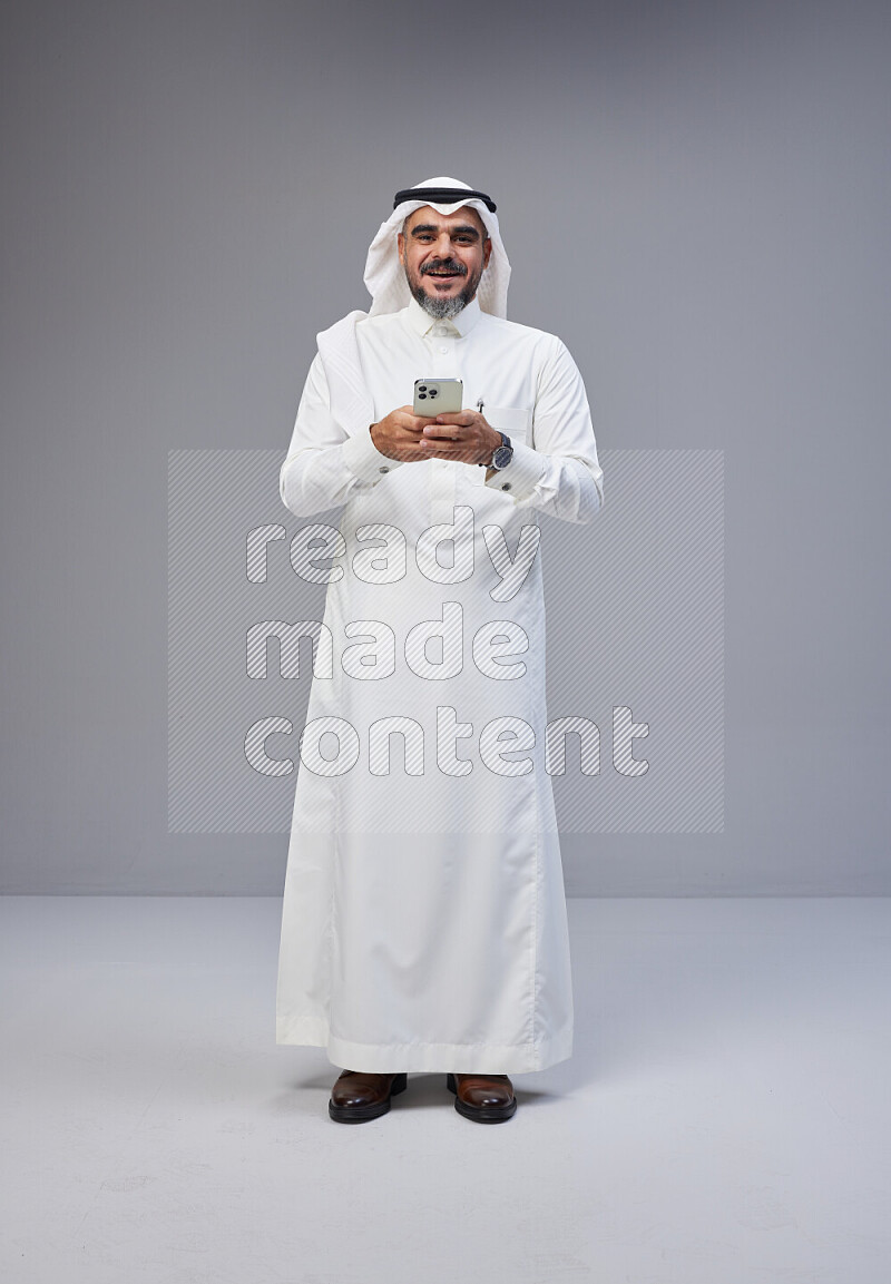 Saudi man Wearing Thob and white Shomag standing texting on phone on Gray background