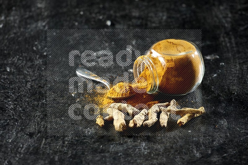 A flipped glass spice jar and metal spoon full of turmeric powder and powder spilled out of it with dried whole fingers on textured black flooring