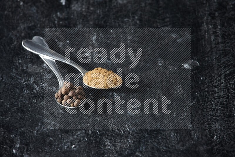 ملعقتان معدنيتان ممتلئتان ببودرة بهار الفلفل الحلو وحبوبه على خلفية سوداء