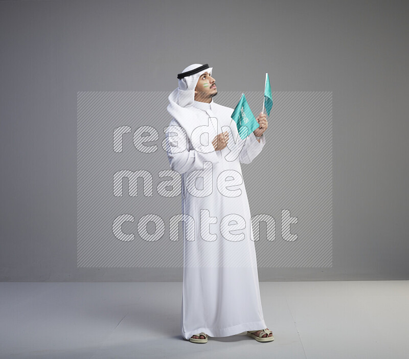 A Saudi man standing wearing thob and white shomag with face painting raising small saudi flag on gray background