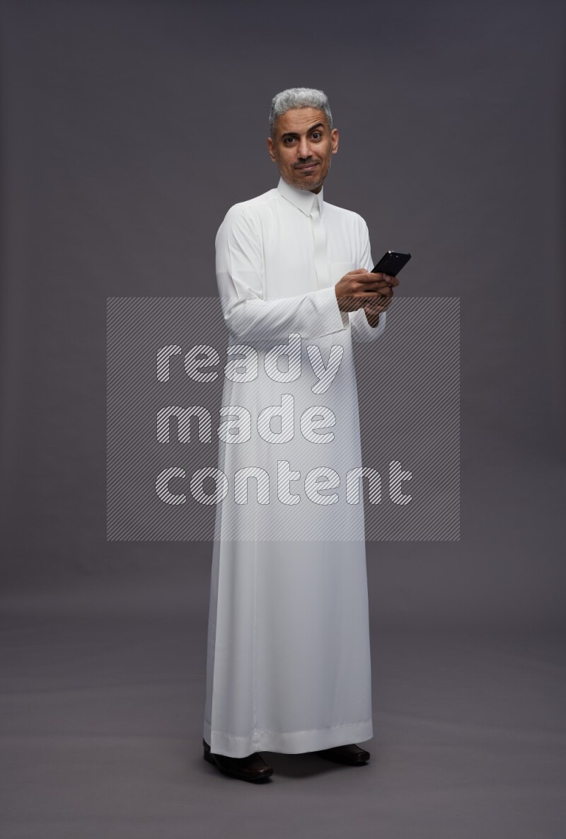 Saudi man wearing thob standing texting on phone on gray background