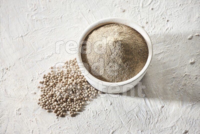 white pottery bowl full of white pepper powder with pepper beads on textured white flooring