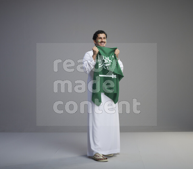 A Saudi man standing wearing thob with face painting wrapping Saudi flag scarf and holding big Saudi flag on gray background