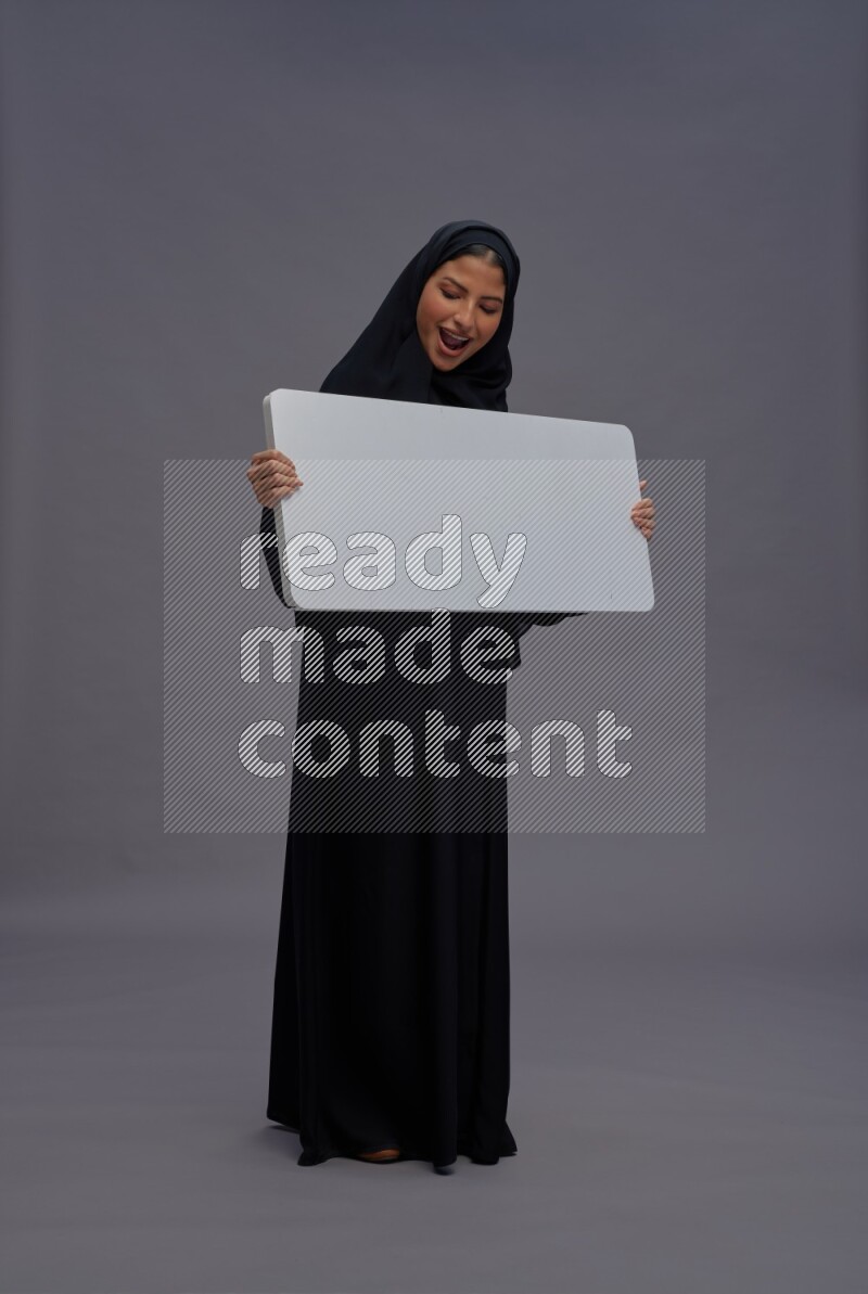 Saudi woman wearing Abaya standing holding white board on gray background