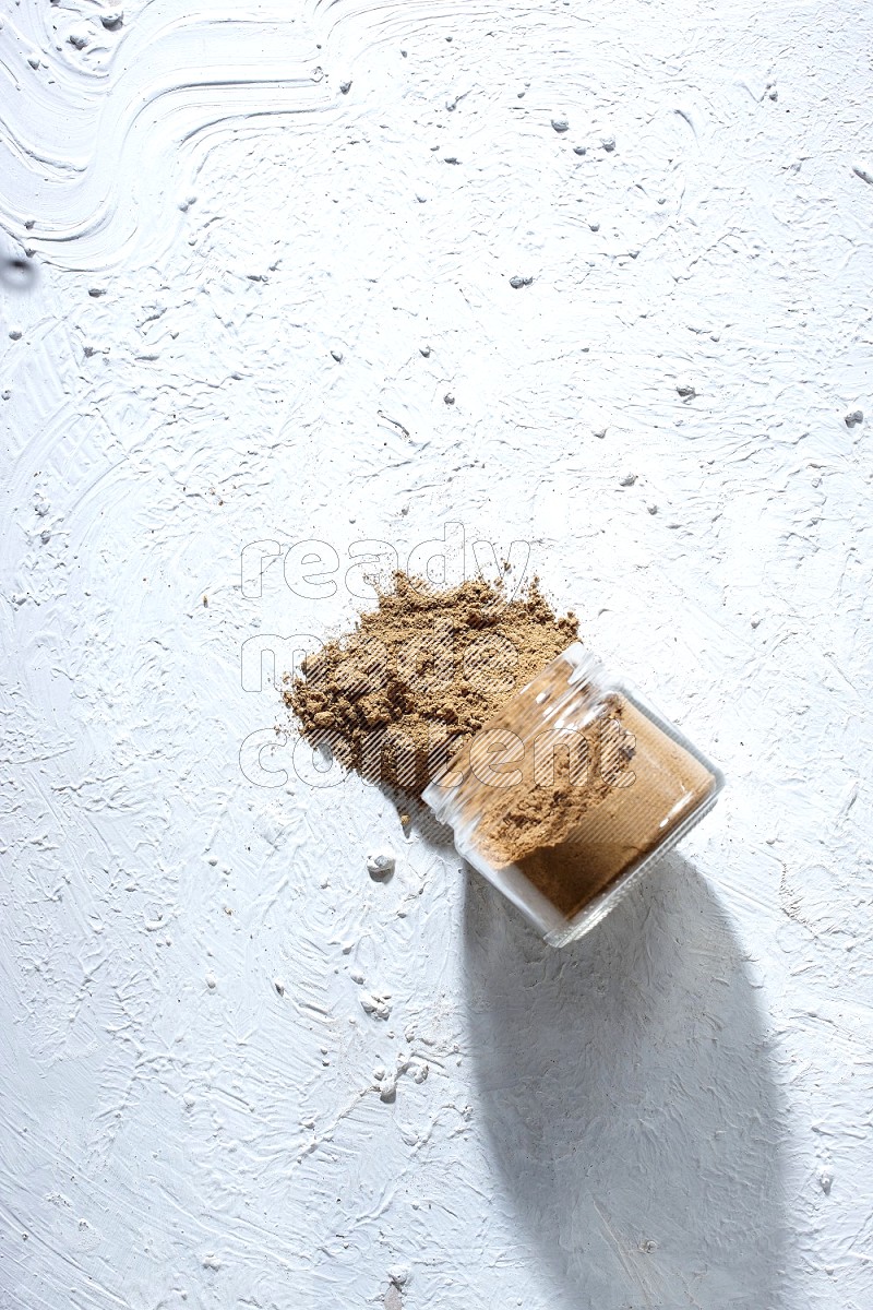 A flipped glass jar full of allspice powder and powder spilled out of it on a textured white flooring