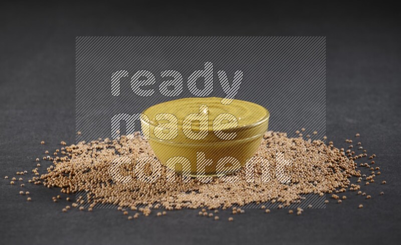 A glass bowl full of mustard paste with mustard seeds underneath on black flooring