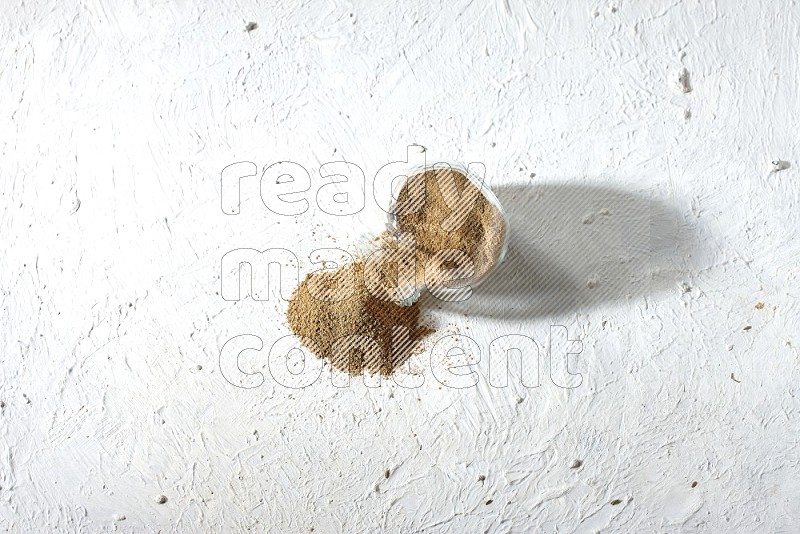 A flipped glass spice jar full of cumin powder and powder spilled out on textured white flooring