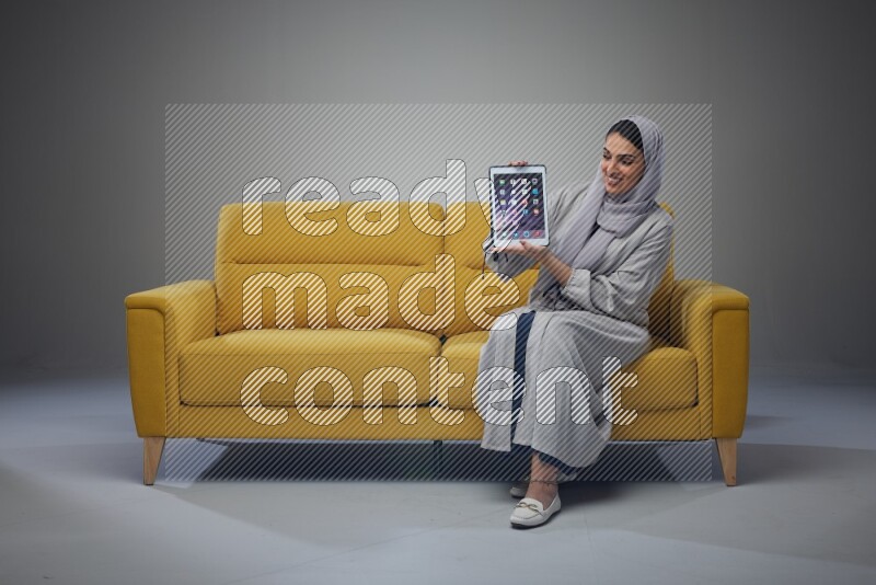 A Saudi female wearing a light gray Abaya and head scarf sitting on a yellow sofa and showing her tablet's screen while wearing headphones eye level on a grey background