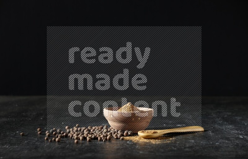 A wooden bowl and spoon full of allspice powder and whole balls spreaded on a black flooring