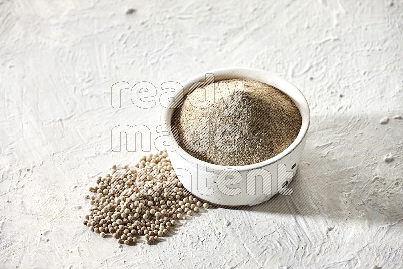 white pottery bowl full of white pepper powder with pepper beads on textured white flooring