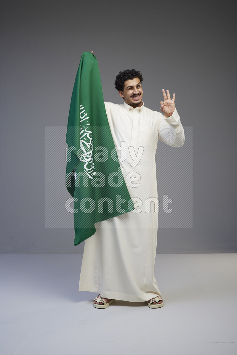 A Saudi man standing wearing thob holding big Saudi flag on gray background