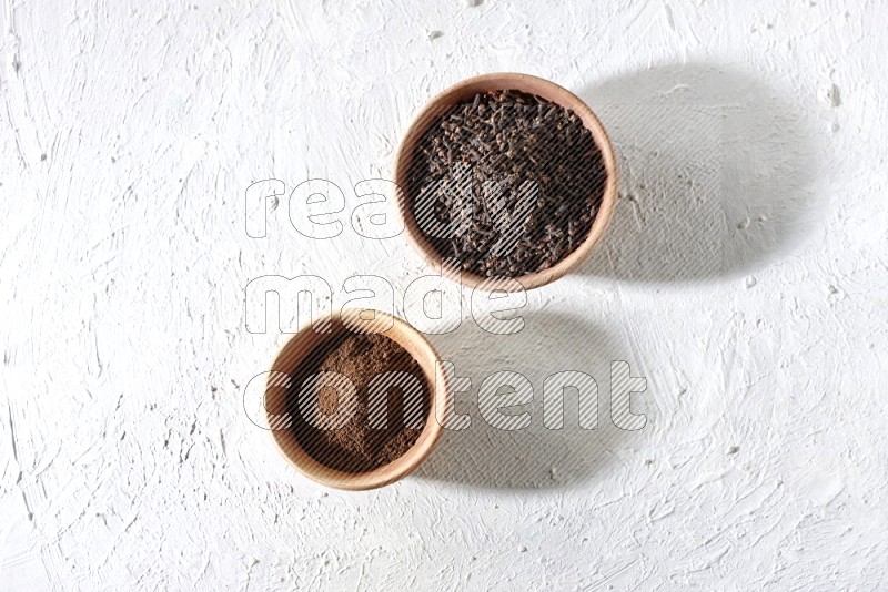 2 wooden bowls full of cloves powder and whole cloves on a textured white flooring