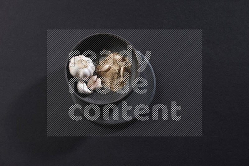 A black pottery bowl full of garlic powder and cloves and a bulb on a black flooring in different angles