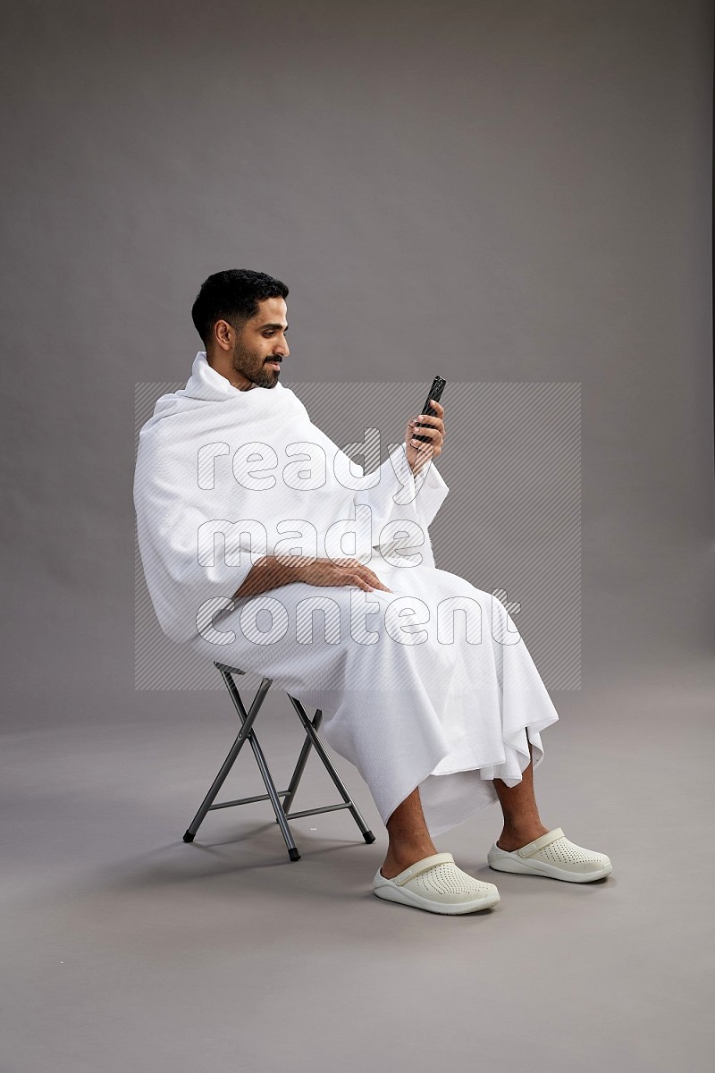 A man wearing Ehram sitting on chair texting on phone on gray background