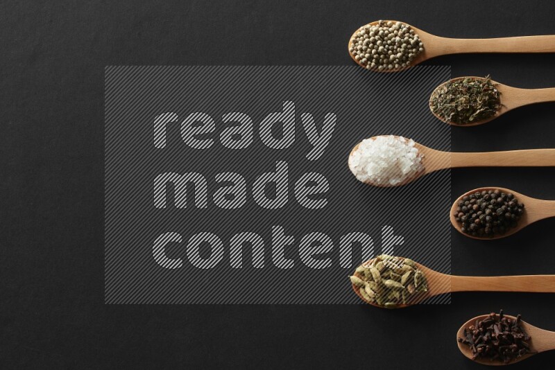 الفلفل الأبيض والقرنفل والهيل والملح والفلفل الأسود وريحان مجفف في معالق خشبية علي خلفية سوداء