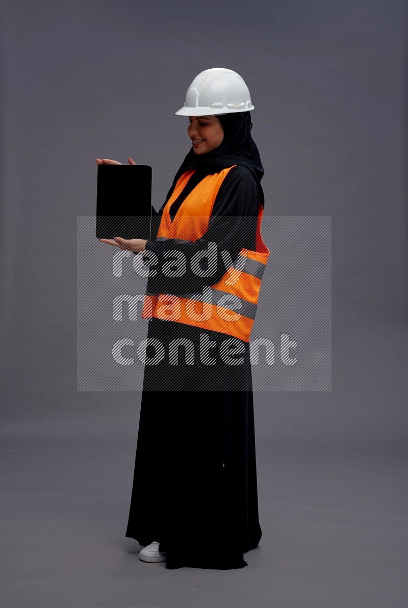 Saudi woman wearing Abaya with engineer vest standing showing tablet to camera on gray background