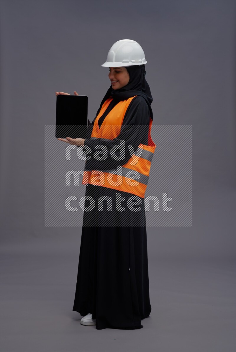 Saudi woman wearing Abaya with engineer vest standing showing tablet to camera on gray background
