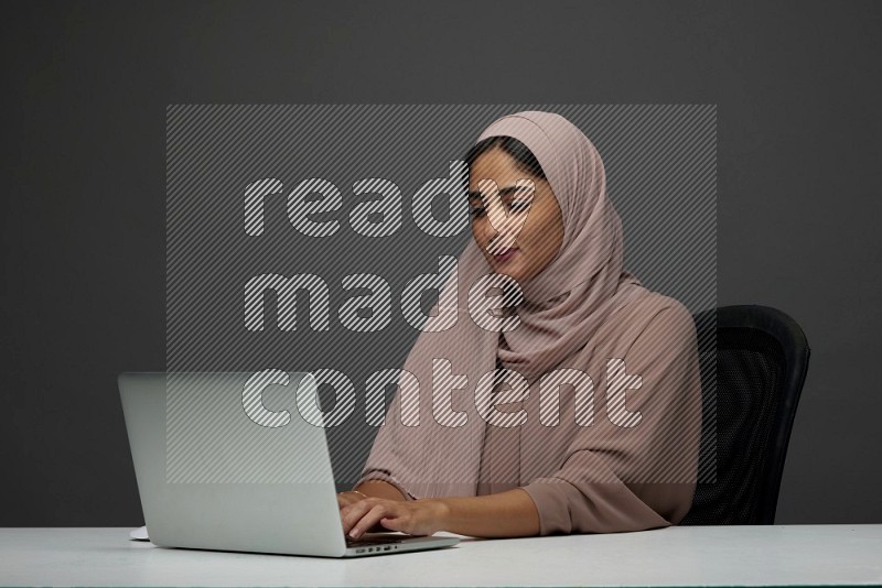 A Saudi woman Setting on her desk Typing on her laptop on a Gray Background wearing Brown Abaya with Hijab