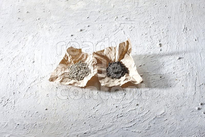 2 Crumpled pieces of paper full of black and white pepper beads on a textured white flooring