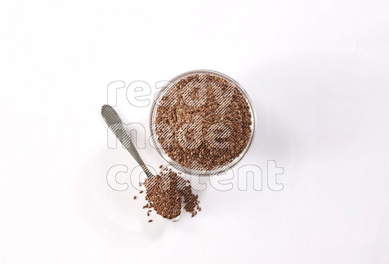 A glass bowl full of flax seeds and a metal spoon full of the seed on a white flooring