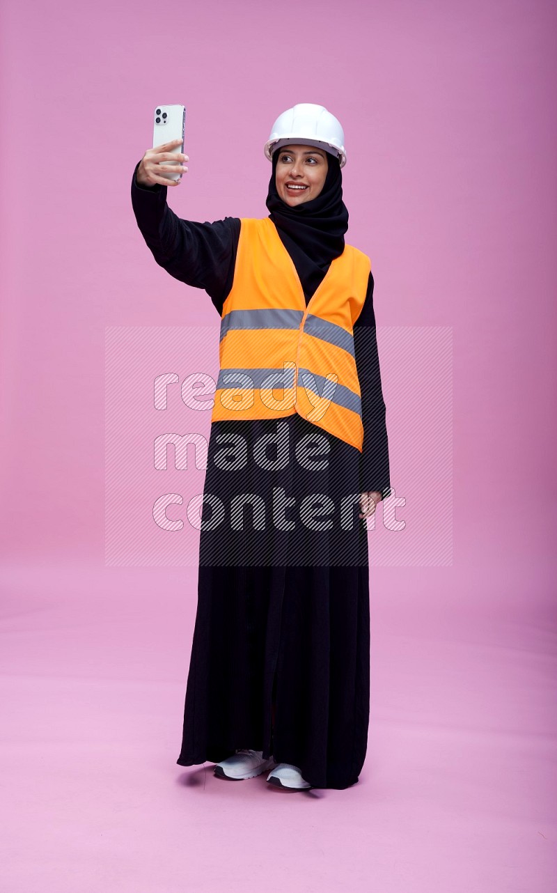 Saudi woman wearing Abaya with engineer vest and helmet standing taking selfie on pink background