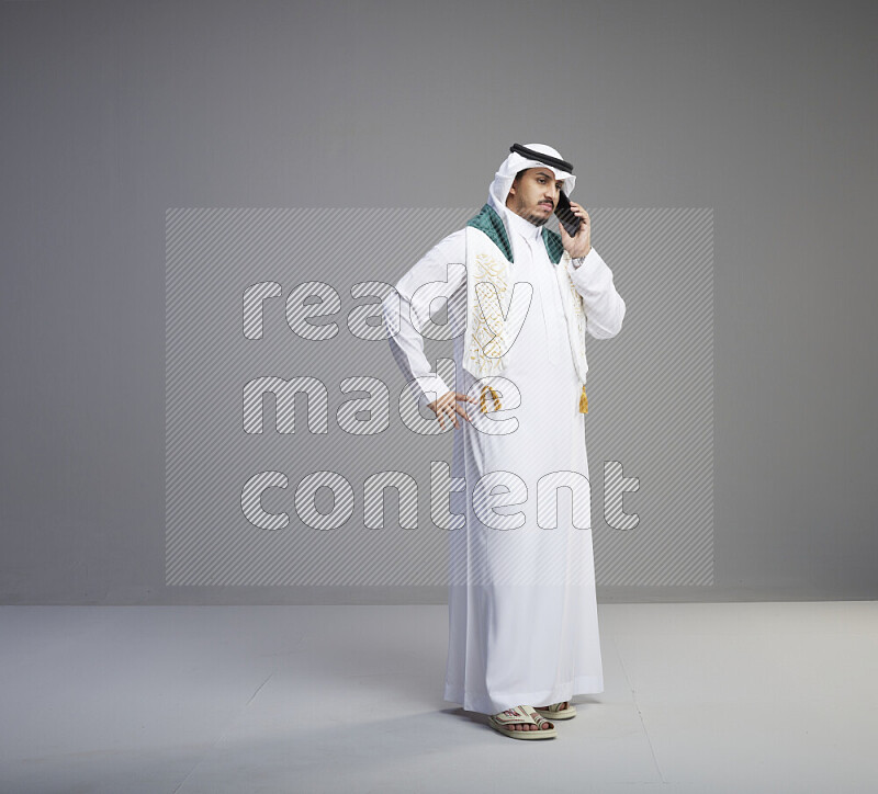 A saudi man standing wearing thob and white shomag with flag scarf on his neck talking on phone on gray background