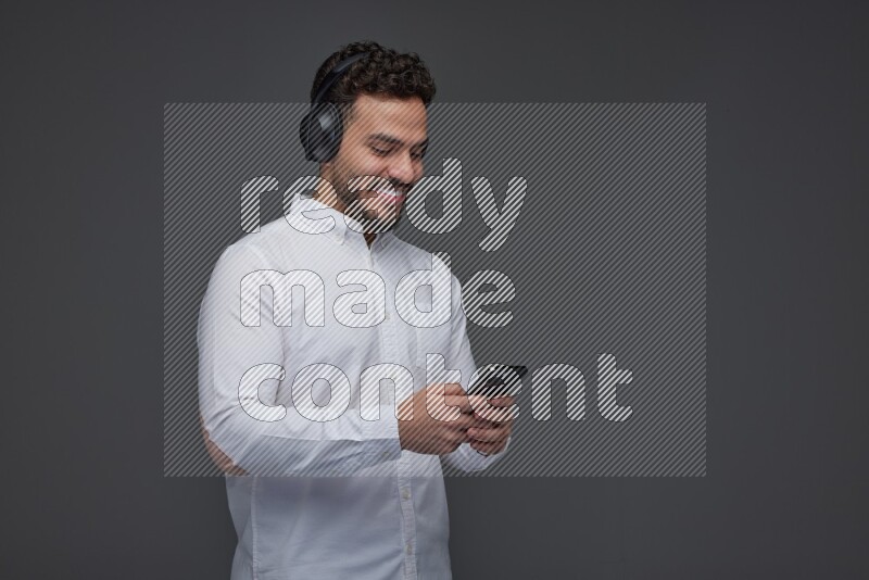 A man wearing smart casual and using his phone and headphone eye level on a gray background