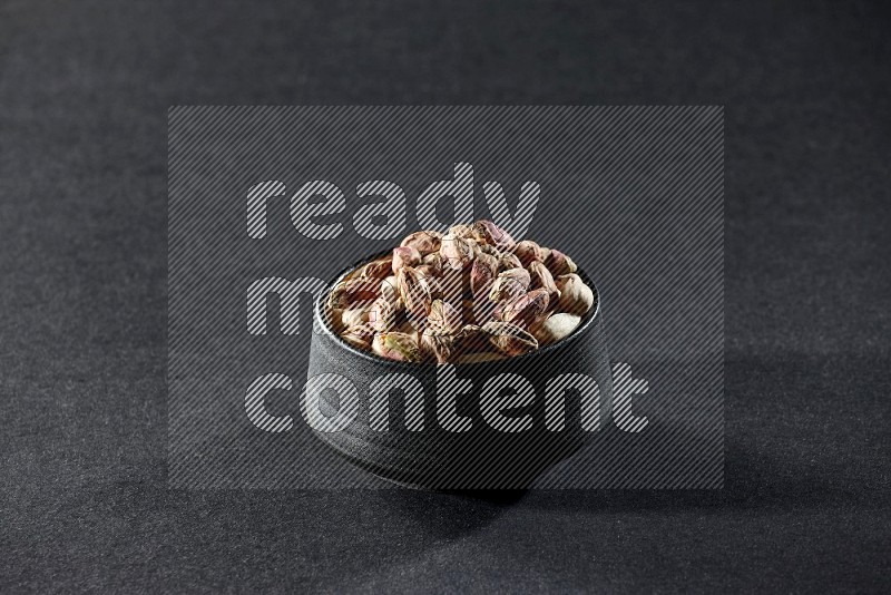 A black pottery bowl full of peeled pistachios on a black background in different angles