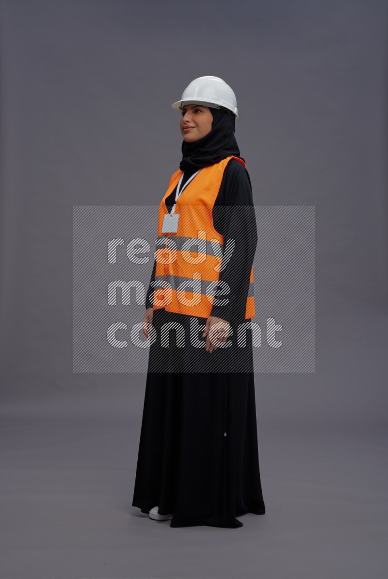 Saudi woman wearing Abaya with engineer vest with neck strap employee badge standing interacting with the camera on gray background