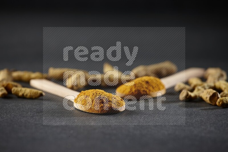 ملعقتان خشبيتان ممتلئتان ببودرة الكركم مع أصابع الكركم المجففة على خلفية سوداء