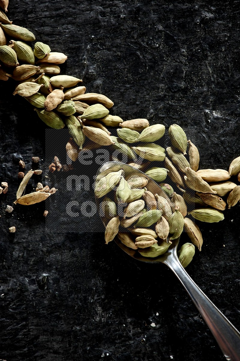 A Metal spoon full of cardamom seeds and some seeds beside it on a textured black flooring