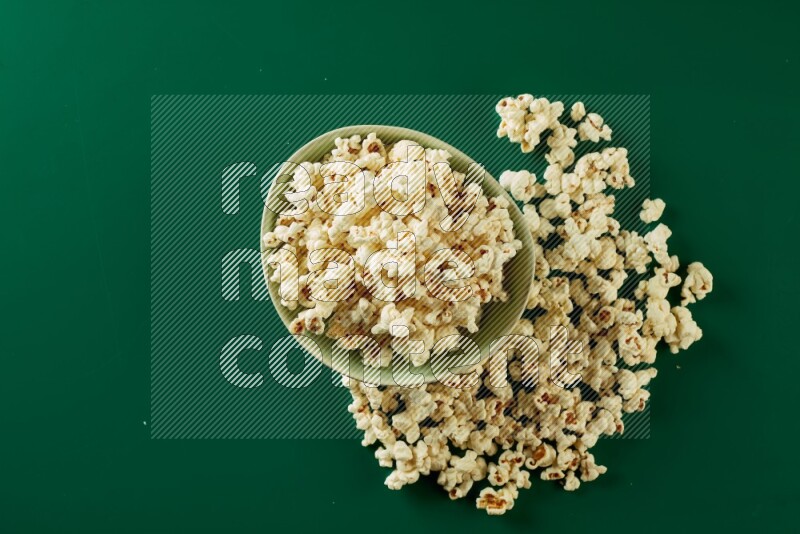 A green ceramic bowl full of popcorn with popcorn beside it on a green background in a top view shot