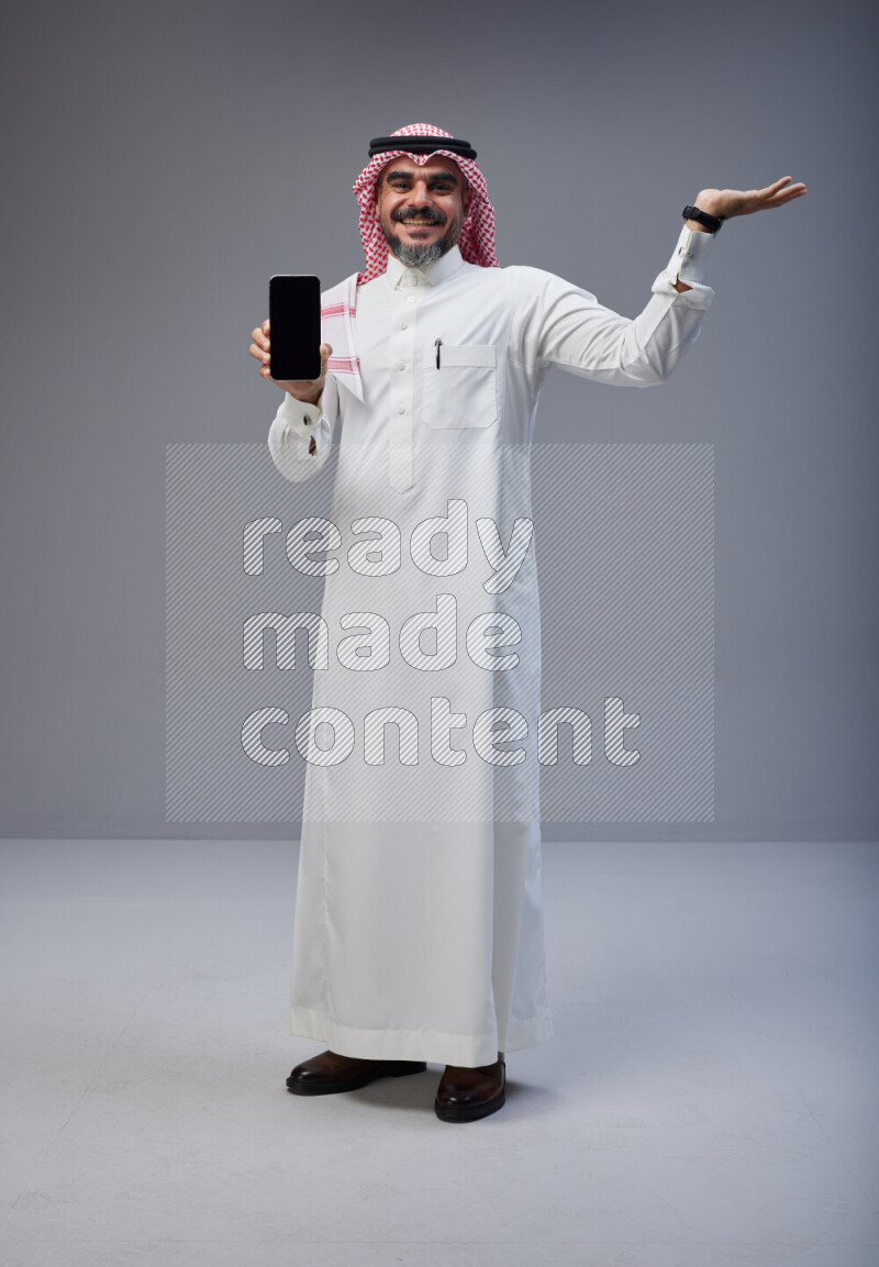 Saudi man Wearing Thob and red Shomag standing showing phone to camera on Gray background