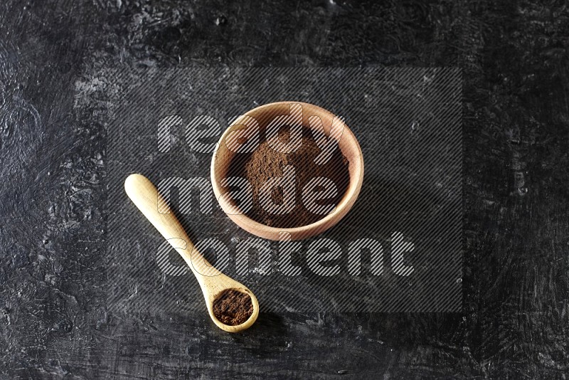 A wooden bowl and wooden spoon full of cloves powder on textured black flooring