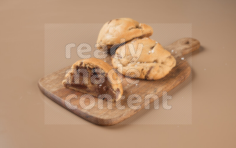 a chocolate chip cookie with another one cut in half on a wooden cutting board on a brown background