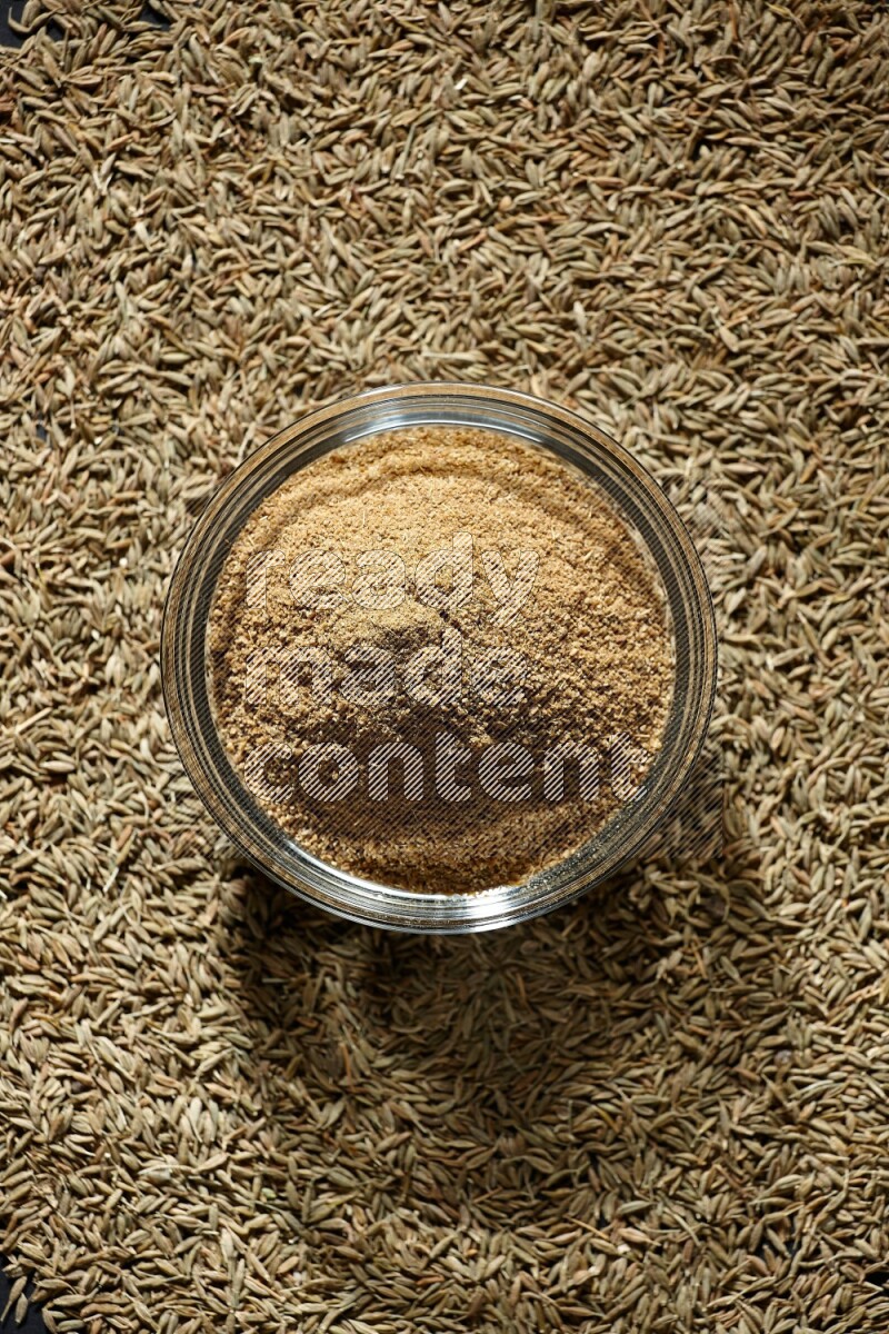 A glass bowl full of cumin powder surrounded by cumin seeds on black flooring